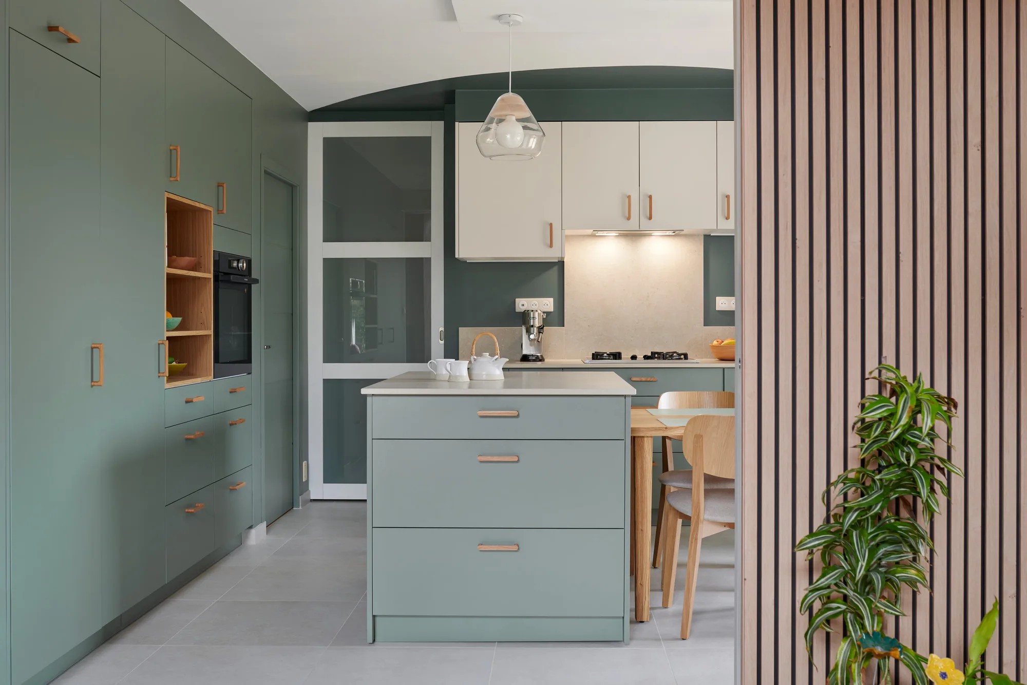 Cuisine ouverte contemporaine aux façades vert eucalyptus mates et poignées bois, îlot central et claustra en tasseaux, photographiée pour un cuisiniste
